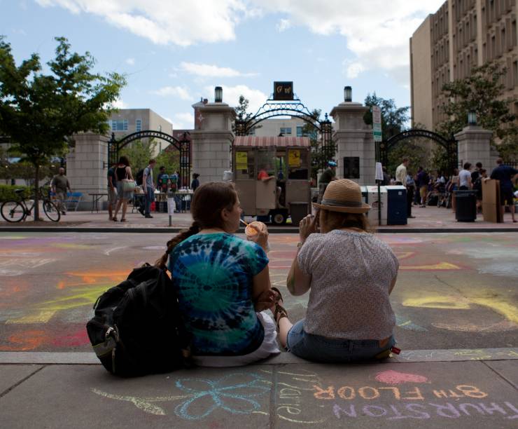 Two students sitting on curb looking at chalk drawings