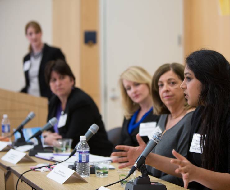 Four women sit on a panel with microphones, listening as one speaks. A fifth woman stands at a podium in the background.