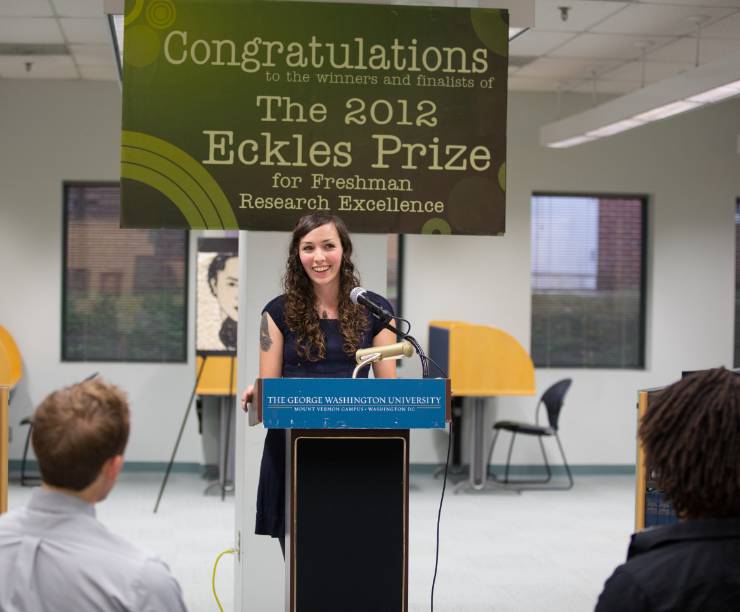 Female student gives speech at a podium in front of a sign that says, 