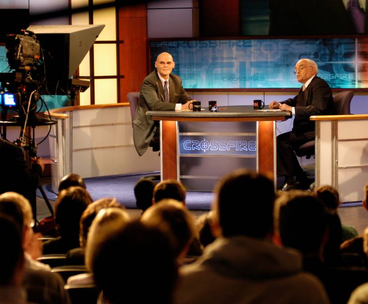 Two hosts sit at a table talking onstage before a full audience. A television camera is visible in the corner.