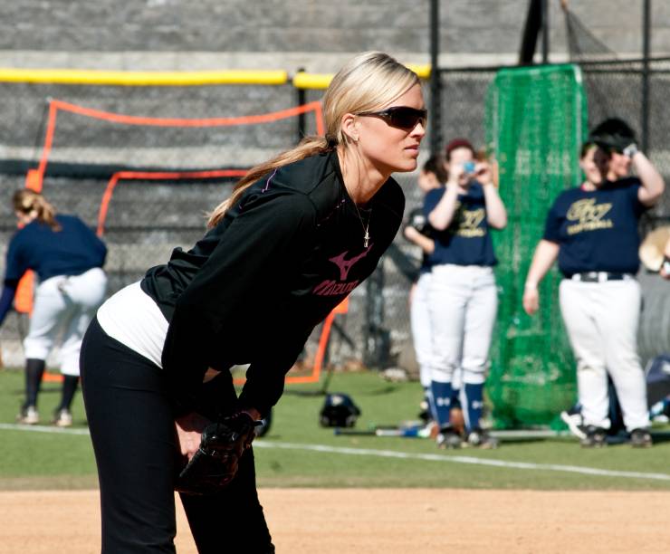 Jennie Finch leans over while players in GW uniforms look on in the background.