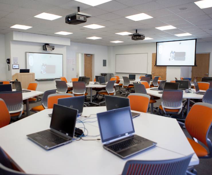 A classroom furnished with laptops on tables and large screens on the walls.