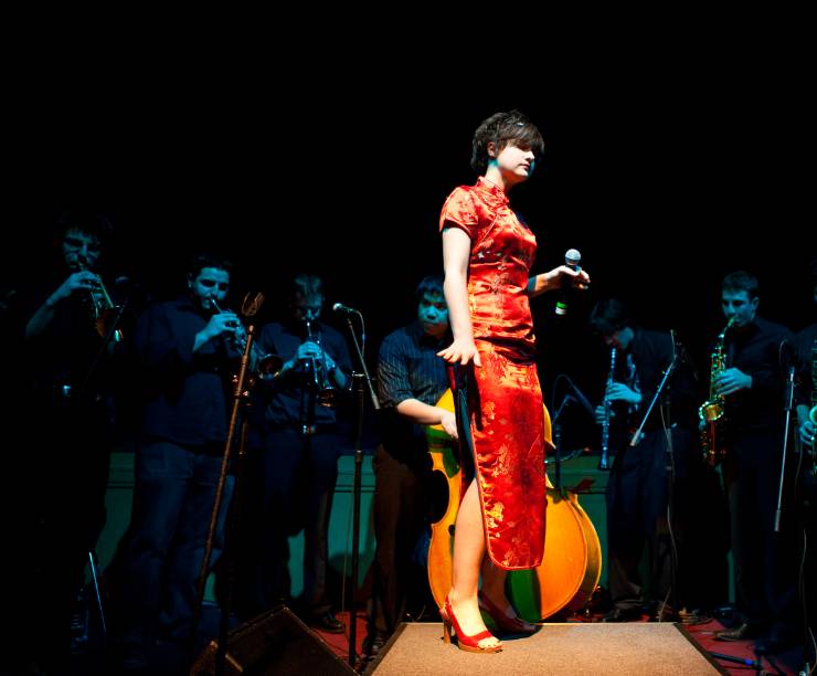 A jazz singer stands in a spotlight surrounded by a group of brass musicians.