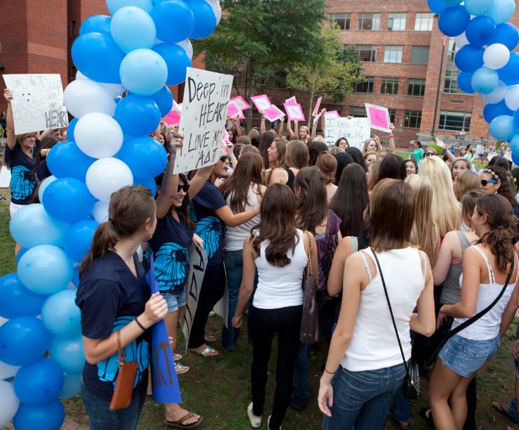 A crowd of students holding signs for various sororities stand under an arc of balloons.