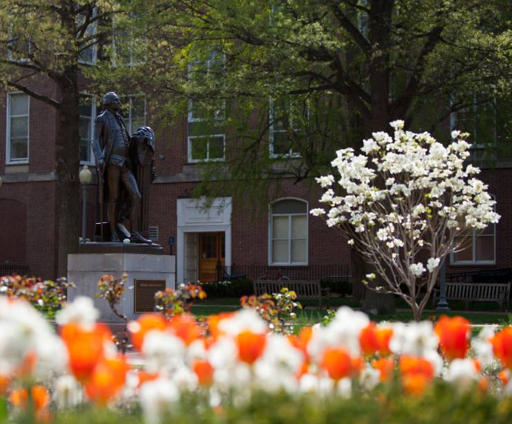 A bronze statue of George Washington stands framed by flowers in the foreground and brick buildings behind.