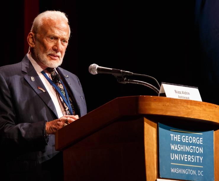 Buzz Aldrin speaks at a podium with a GW logo