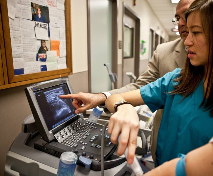 Two female students wearing scrubs observe a sonogram image with an instructor.