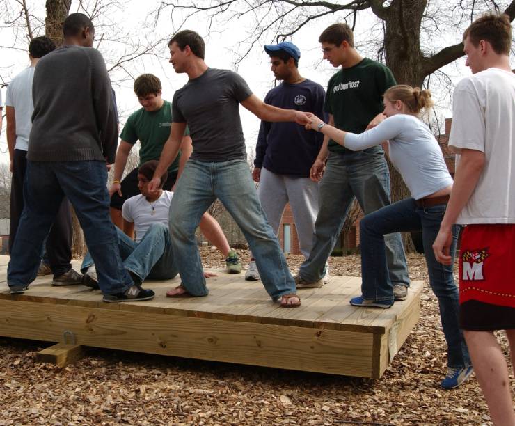 A group of participants is standing on a wooden platform and trying to balance their weight as they help another participant join them on the platform.