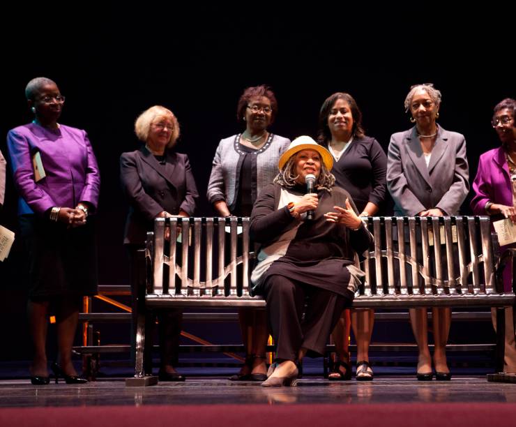 Toni Morrison sits speaking on a bench onstage with a group of women standing behind her.