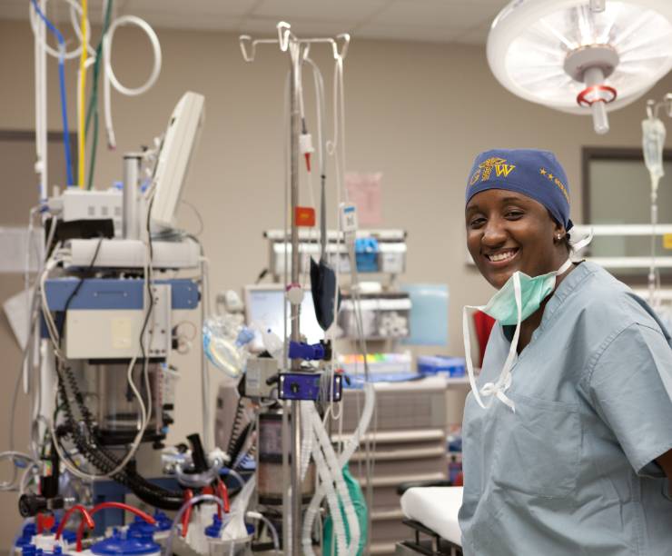 Female doctor wearing scrubs and a mask stands in an operating room with medical equipment.