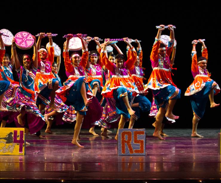 A group of students in traditional brightly-colored costumes dance onstage.
