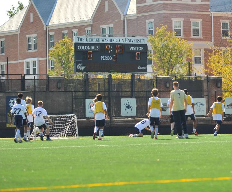 Kids play soccer on a field supervised by adults. Brick buildings are in the background.
