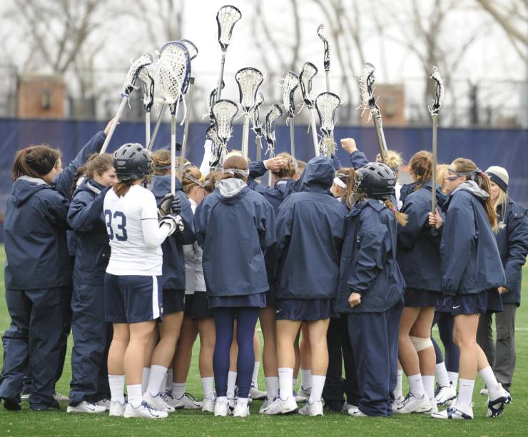 GW Women's Lacrosse team stands in a huddle with their sticks raised before a game.