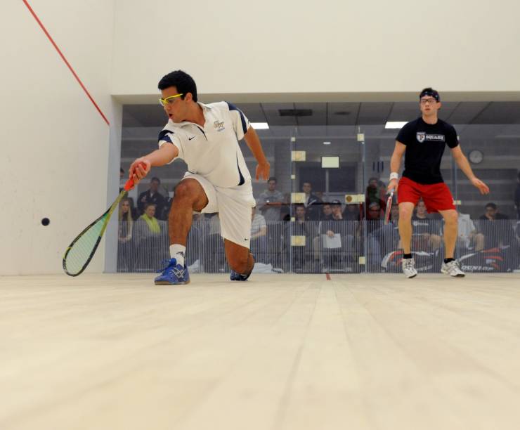 Two squash players play while spectators watch from outside the court.