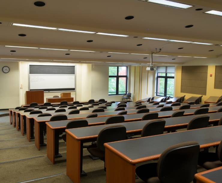 Classroom shot of GW Law's Lerner 201. The classroom includes long desks with chairs and a whiteboard at the front of the classroom. Windows featured in the front of the classroom look out to University Yard.