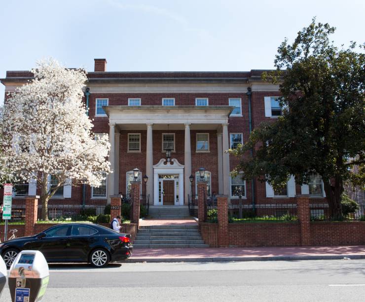 Red brick building with four white columns, stairs and a blooming tress.