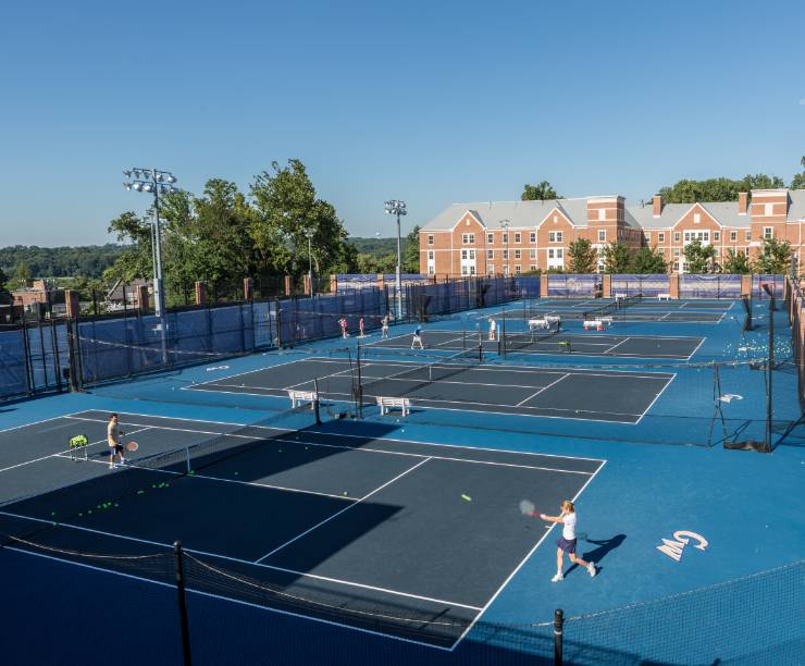 Blue outdoor tennis courts with a few students practicing on them.