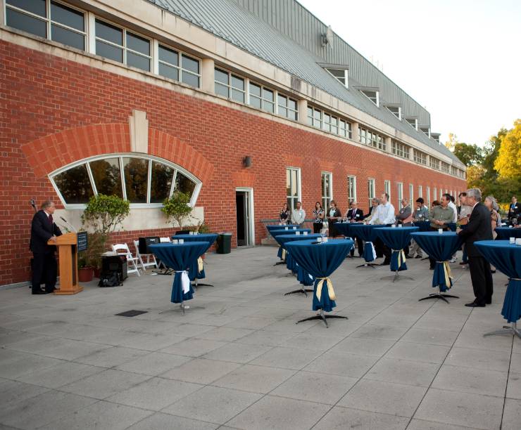 People stand on a terrace at tableclothed cocktail tables and listen to a speaker.