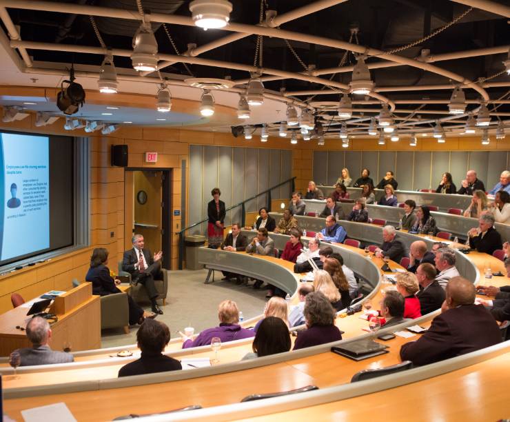 People sit in tiered seating around a lecture hall listening to two speakers.