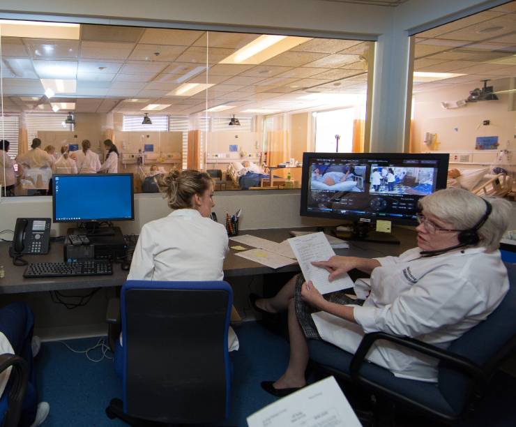A professor and students sit in a control room at computers, looking out of one-way mirrors at a simulated hospital floor where students are with a mannequin patient.