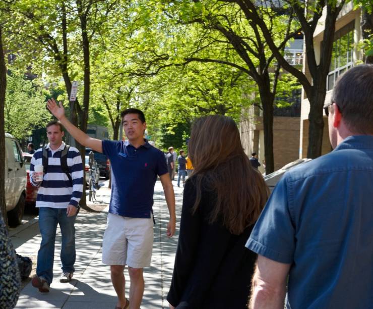 Tour leader leads a group of visitors visiting campus. The group is walking down the street and the tour leader is pointing up.