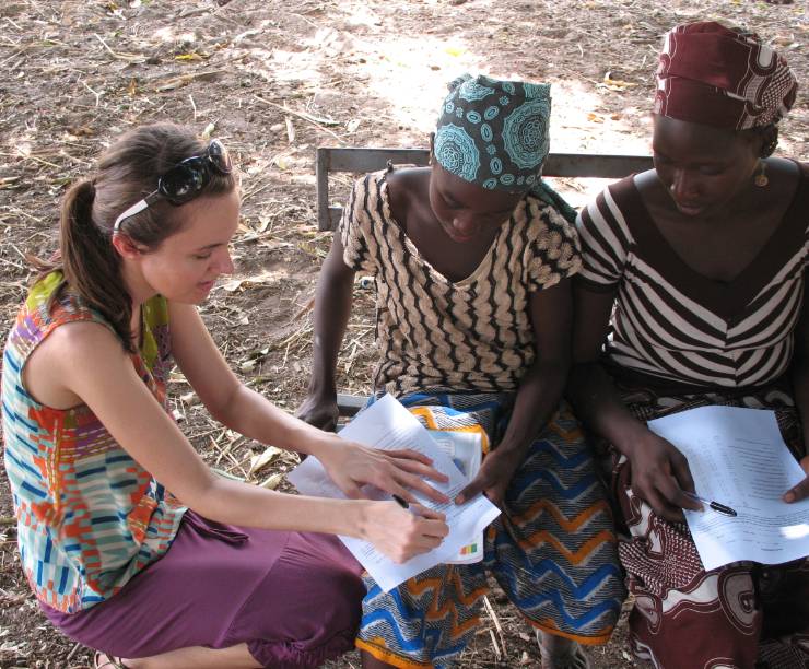 GW student visits with two women in Mali. The three woman are looking down at a piece of paper the student is writing on.