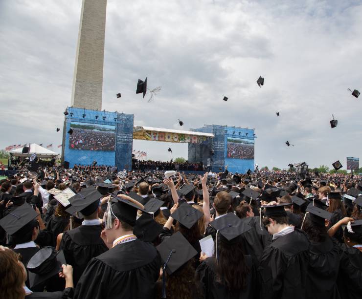 Photo of outdoor commencement ceremonies showing thousands of graduates sitting in cap and gown facing a stage with the washington monument in the background.