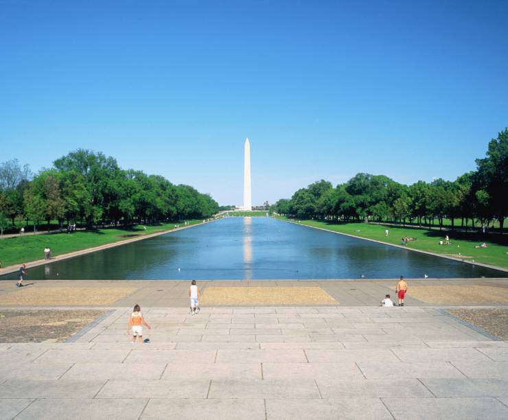 Photo with long rectangular pool of water, green grass, a blue sky and the washington monument in the background.