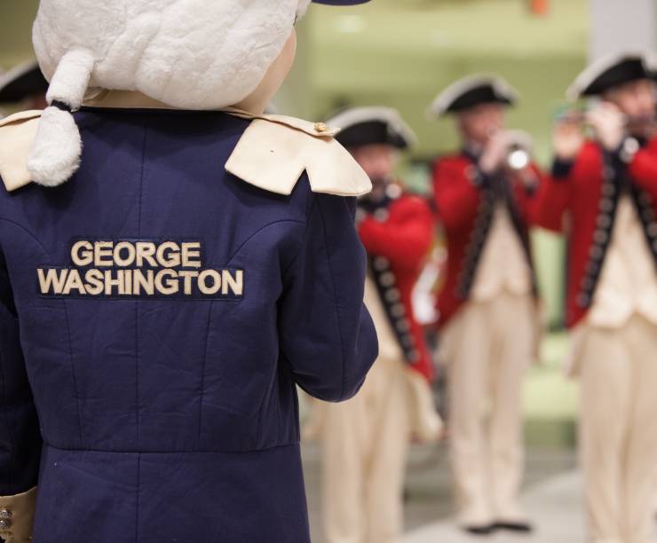 Mascot George stands facing men in colonial uniforms.