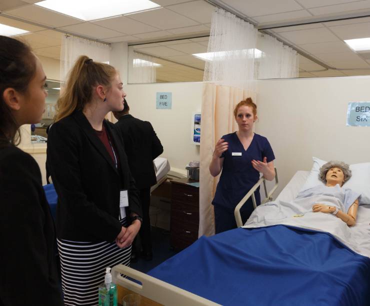 Nursing student is standing next to a simulation mannequin while addressing a group of high school students.