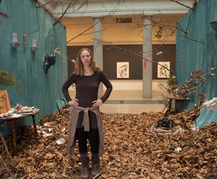 A woman stands in an art installation - with dry leaves on the floor and twigs and sconces attached to blue-painted walls.