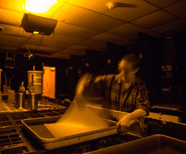 A student pulls a piece of photographic paper from a developer bath in a dark room.