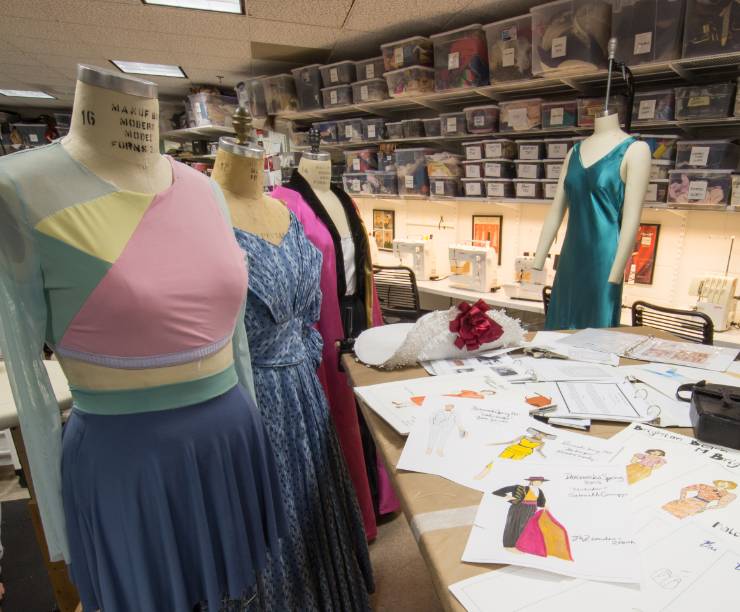 The inside of a costume design workshop features dress forms with dresses under construction and costume sketches on a table.