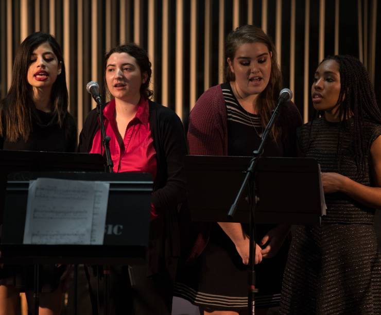 Four women stand behind music stands and microphones and sing.