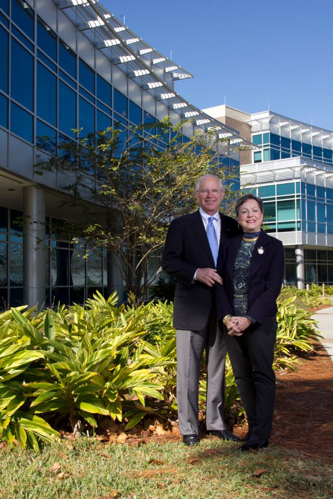 An older Caucasian male and female stand in front of a two-story glass fronted building with green foliage on a bright autumn day.